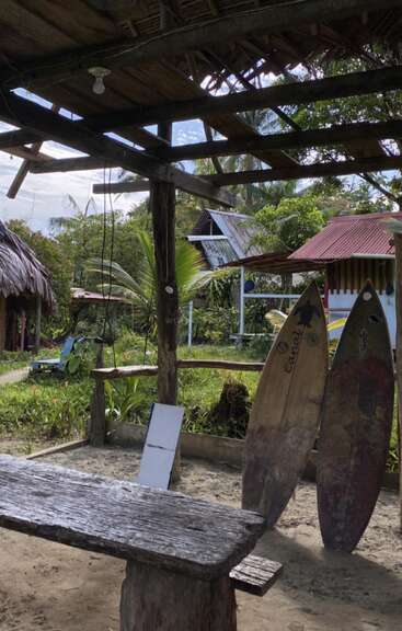 Dans cette scène rustique en plein air, des bancs et une table en bois sont placés sous un toit de chaume. Deux planches de surf usées reposent à proximité, entourées de plantes tropicales et de huttes.