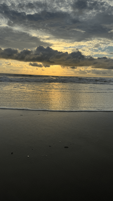 A luz dourada do sol atravessa nuvens dramáticas, refletindo na areia molhada e nas ondas suaves ao longo de uma praia tranquila, evocando calma e beleza natural na cena serena.