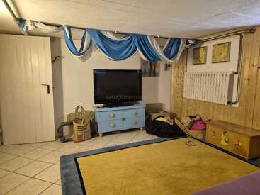 A cozy basement room with a TV on a blue dresser, yellow rug, patterned ceiling drapes, wooden chest, baskets, radiant heater, and tiled floor.