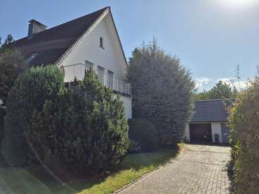 A white house with a steep roof is surrounded by lush greenery and trees. A paved driveway leads to a detached garage under a bright sunny sky.