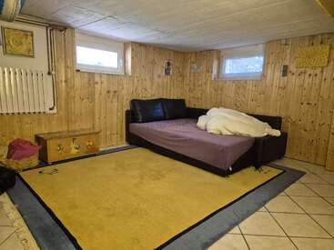 This cozy basement room features wood-paneled walls, a sofa bed with rumpled bedding, small windows, children’s chest, playful yellow rug, and a tiled floor.