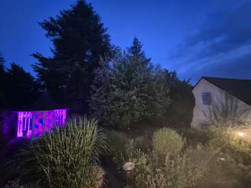 A peaceful garden at dusk features lush greenery, a gazebo glowing with purple lights, a house with blue shutters, and soft outdoor lighting, creating a serene atmosphere.