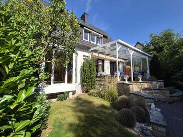 A charming house with a sloped roof, large windows, and a glass-covered patio, surrounded by lush greenery, shrubs, lawn, and stone terrace under clear skies.
