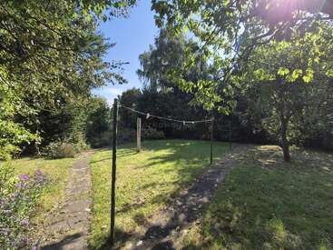 This peaceful garden features lush green grass, trees casting gentle shadows, a stone path curving through, and a clothesline with colorful pegs illuminated by sunlight.