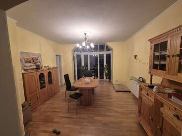 A cozy dining room with wooden furniture, yellow walls, and hardwood floors. Central table, chairs, plants by large windows, a chandelier overhead, and decorative items throughout.