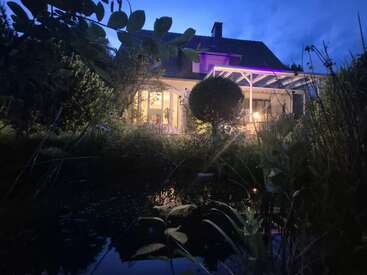 A cozy house glows warmly in the evening, surrounded by lush greenery and a tranquil pond, with vibrant lights illuminating the patio under a twilight sky.