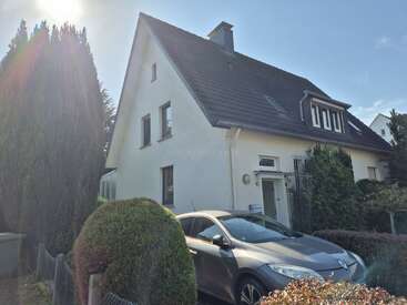 A modern white house with a steep black roof, large windows, and a front garden. A silver car is parked in the driveway, surrounded by greenery.