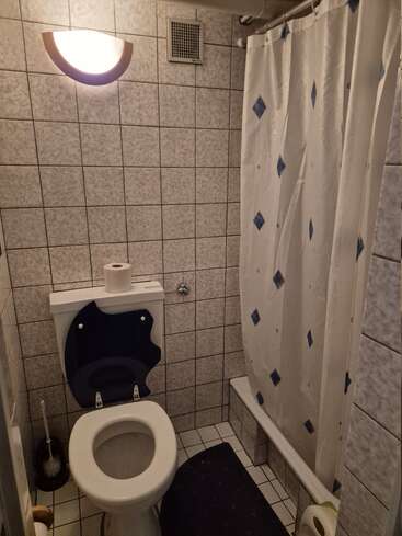This is a small bathroom with tiled walls, a toilet, toilet brush, toilet paper, a shower with a curtain, a wall light, and a black bath mat.