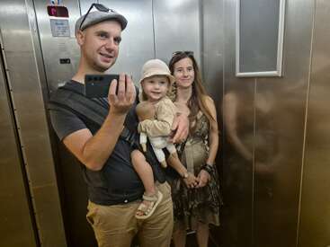 A family of three takes a selfie in an elevator. The father holds a phone and carries a child, while the mother stands beside them, smiling.