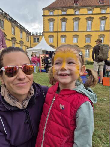 A smiling woman in sunglasses poses with a cheerful young girl, whose face is painted like a lion, at an outdoor event near a yellow building.