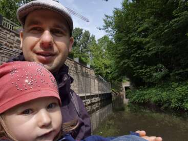 A man wearing a cap and a child in a red hat pose for a selfie near a canal, surrounded by lush greenery and stone walls.