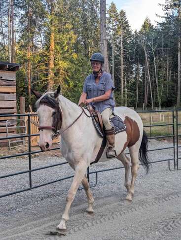 A imagem mostra uma mulher montada em um cavalo branco com manchas marrons, usando um capacete e uma camisa azul, em uma área cercada, rodeada por árvores e uma estrutura de madeira.