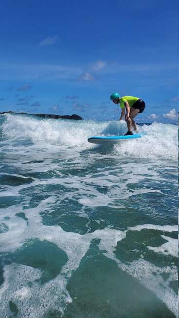 Uma pessoa com camisa verde e capacete surfa uma onda em uma prancha de surfe azul sob um céu claro e ensolarado com água espumosa e terra distante.