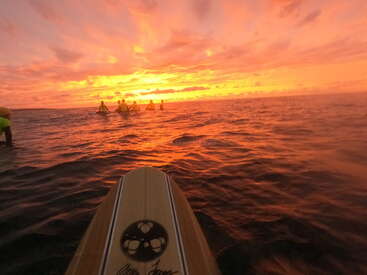 Uma prancha de surfe de madeira aponta para um grupo de surfistas sentados no oceano brilhante, banhado por um incrível e vibrante céu laranja e amarelo ao pôr do sol.