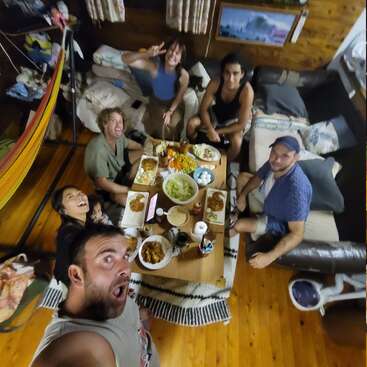 Six friends gather around a table full of delicious food, smiling and posing for a selfie in a cozy, wooden room with a hammock and cushions.