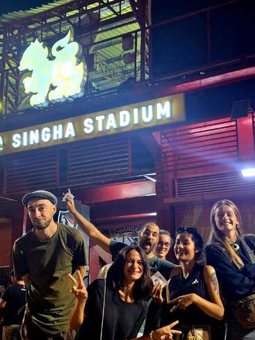 A group of six smiling friends pose outside Singha Stadium at night, flashing peace signs. Above them, a glowing Singha lion logo and stadium sign are visible.
