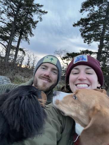 Ein lächelndes Paar mit Mützen macht ein fröhliches Selfie im Freien mit zwei Hunden. Hohe Bäume und ein wolkenverhangener Himmel bilden eine friedliche, natürliche Kulisse, die das Glück einfängt.
