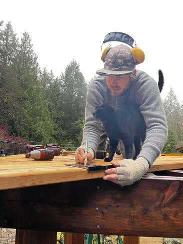 Ein Mann mit Gehörschutz und Handschuhen markiert Holz mit einem Bleistift, während eine schwarze Katze neben ihm steht; Werkzeuge sind auf der Holzoberfläche verstreut. Draußen.
