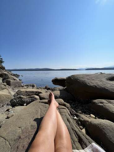 A person relaxes on rocky shore, legs outstretched, looking towards calm water, distant forested hills, and clear blue sky on a sunny, peaceful day.