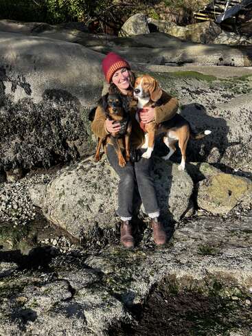 A smiling person wearing a red beanie and brown jacket sits on a rocky surface, happily hugging two dogs—one brown and black, the other brown and white.