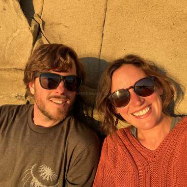 A smiling man and woman wearing sunglasses, sitting together in warm sunlight against a textured stone wall, enjoying a relaxed moment and casting gentle shadows around them.