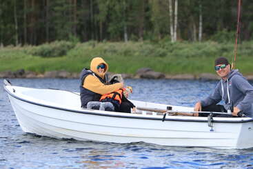 Dos personas con sudaderas con capucha y gafas de sol están sentadas, sonriendo, en una pequeña barca blanca en un lago, acompañadas por un perro que lleva un chaleco salvavidas naranja. Fondo de bosque.