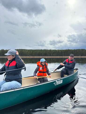 Trois personnes, vêtues de gilets de sauvetage et coiffées de chapeaux, pagayent dans un canoë vert baptisé « Pelican » sur un lac calme, sous un ciel nuageux, au milieu d'une forêt.