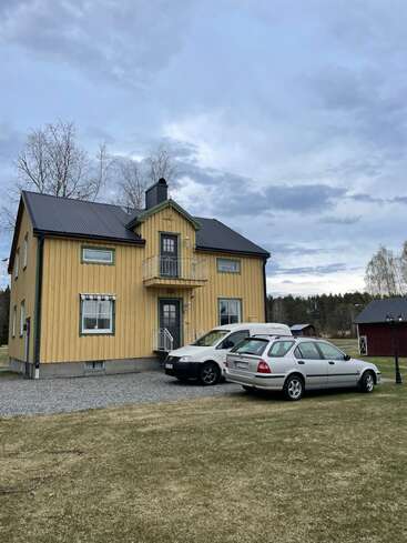 Une maison jaune à deux étages, au toit noir, se dresse sur un terrain gazonné. On aperçoit deux voitures garées. Le ciel est nuageux et des arbres entourent les lieux.