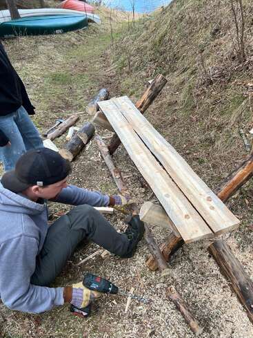 Un homme est en train de construire un banc rustique en bois à l'extérieur, à l'aide de rondins et de planches. Il tient une perceuse, tandis qu'une autre personne se tient à proximité. Des canoës sont visibles à l'arrière-plan.