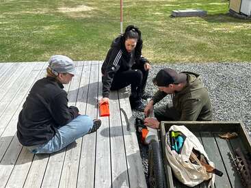 Trois personnes sont assises dehors sur une terrasse en bois et réparent un vélo. Des outils et des pièces sont éparpillés tout autour. Elles travaillent ensemble, concentrées et absorbées par leur tâche, sous un ciel nuageux.