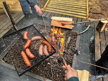 Plusieurs saucisses cuisent sur un feu de camp, tandis que des personnes tiennent des brochettes pour faire griller d'autres saucisses. La scène se déroule en plein air, sur une terrasse en bois.