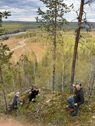Sur une colline recouverte de mousse, trois personnes contemplent une route sinueuse, une rivière et un vaste paysage forestier sous un ciel nuageux. De grands arbres encadrent cette vue panoramique.