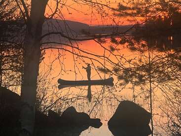 Une personne solitaire se tient debout dans un canoë sur un lac calme au coucher du soleil, entourée de silhouettes d'arbres et de rochers, créant une atmosphère paisible et sereine.