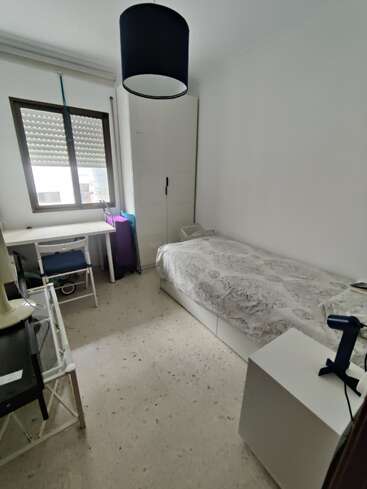 Das kleine, aufgeräumte Schlafzimmer verfügt über ein Einzelbett, einen weißen Kleiderschrank, einen Schreibtisch mit Stuhl, einen Nachttisch, eine Lampe und ein großes Fenster, durch das Tageslicht fällt.