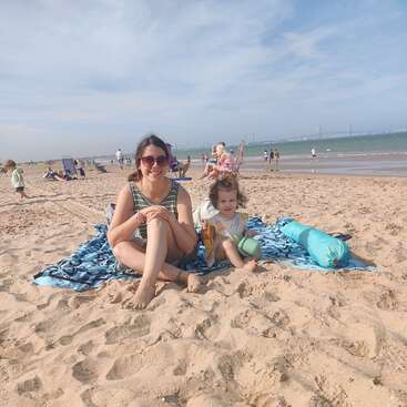 Eine Frau und ein kleines Kind sitzen auf einer Decke an einem Sandstrand und genießen einen sonnigen Tag. Im Hintergrund sind Menschen und das Meer zu sehen.