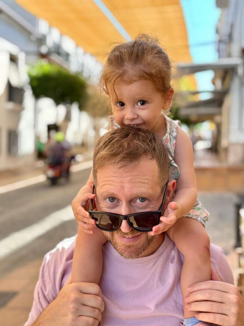 Ein Mann mit Sonnenbrille lächelt, während er ein junges Mädchen auf seinen Schultern trägt. Das Mädchen beißt ihm spielerisch in den Kopf. Sie sind an einem sonnigen Tag im Freien.