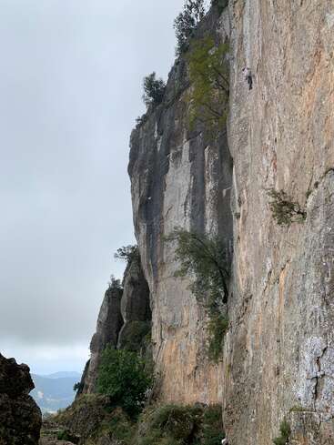 Eine Person klettert an einer hohen, zerklüfteten Felswand mit spärlicher Vegetation. Der Himmel ist bewölkt, und im Hintergrund sind ferne Berge zu sehen, was eine dramatische Szenerie ergibt.