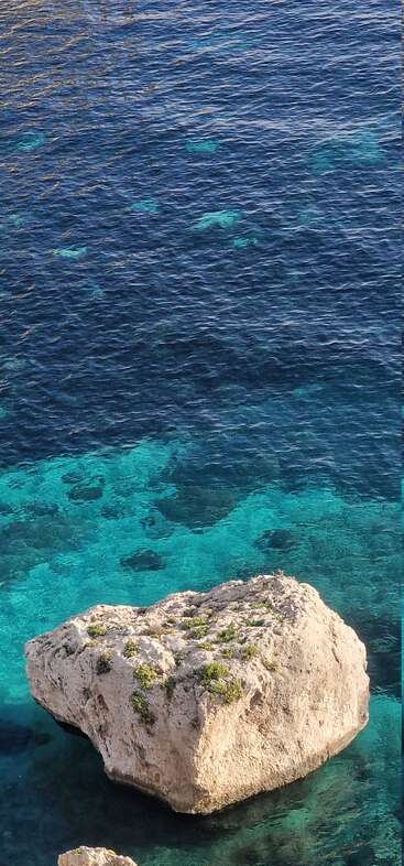 Ein großer, schroffer Felsen ragt aus dem seichten, kristallklaren, türkisfarbenen Wasser empor, während sich im Hintergrund das tiefere, dunkelblaue Meer friedlich ausbreitet.