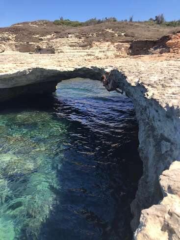 Jemand klettert unter einem natürlichen Felsbogen hindurch, der über klarem, blauem Wasser thront. Um ihn herum erstreckt sich eine raue Küstenlandschaft mit spärlicher Vegetation unter einem strahlend blauen, wolkenlosen Himmel.