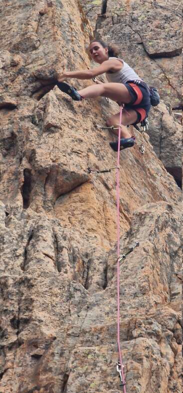 Eine Frau klettert an einer zerklüfteten Felswand empor, gesichert durch ein rosa Seil und einen Klettergurt, und zeigt dabei Entschlossenheit, Kraft und Konzentration bei einem Kletterabenteuer in der freien Natur.