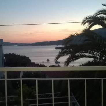 L'image représente une scène sereine de coucher de soleil depuis un balcon, avec un plan d'eau tranquille, des bateaux, des palmiers et des montagnes lointaines, le tout dans un ciel éclatant.