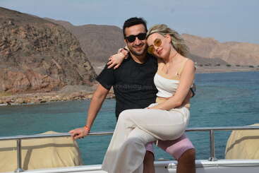 Um casal posa alegremente em um barco, com montanhas rochosas deslumbrantes e águas azuis cristalinas ao fundo; os dois estão usando óculos escuros e roupas leves e de verão.