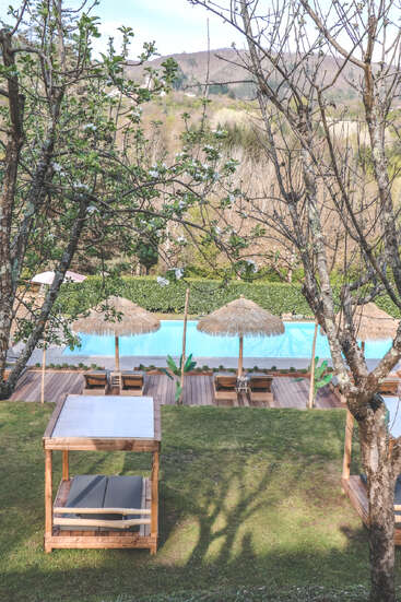 Ein ruhiger Blick auf den Pool, umgeben von Bäumen, Holzhütten, Liegestühlen und strohgedeckten Sonnenschirmen vor der Kulisse üppig grüner Hügel und der Natur.