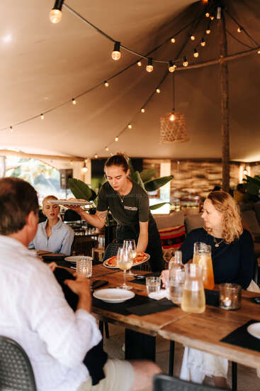 Eine Kellnerin serviert drei Personen in einem gemütlichen, warm beleuchteten Restaurant eine Pizza. Die Gruppe sitzt an einem Holztisch mit Getränken, unter Lichterketten.