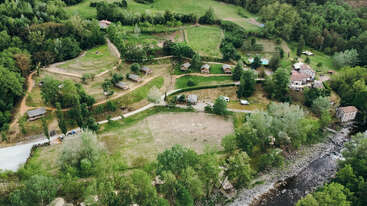 Dieses Luftbild zeigt eine ländliche Szene mit kleinen Hütten, grünen Feldern, gewundenen Wegen, Häusern, einem Fluss und dichten Bäumen in der Umgebung, die eine friedliche Atmosphäre schaffen.