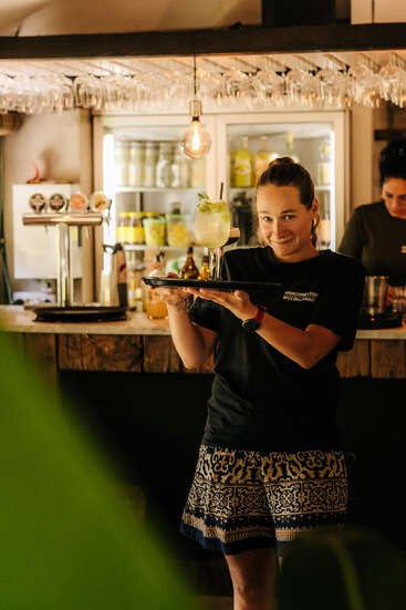 Eine lächelnde Frau steht in einer Bar und hält ein Tablett mit einem ausgefallenen Cocktail. Hinter ihr stehen Flaschen und Gläser. Eine andere Person arbeitet im Hintergrund.