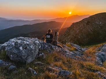 Ein Mensch und zwei Hunde sitzen auf felsigem Gelände und beobachten friedlich einen atemberaubenden Sonnenuntergang über sanften Hügeln und Bergen, umgeben von Gras und natürlicher Schönheit.