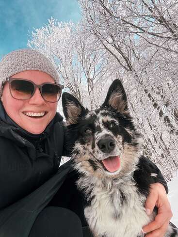 Eine lächelnde Person mit Sonnenbrille und Strickmütze posiert mit einem glücklichen, flauschigen schwarz-weißen Hund. Schneebedeckte Bäume und ein strahlend blauer Himmel umgeben sie.