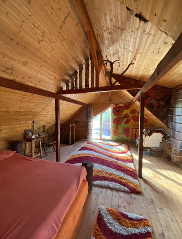Este acogedor dormitorio abuhardillado cuenta con cálidas paredes y techo de madera, una colorida alfombra de rayas, una cama naranja, un pequeño escritorio, una decoración única y luz natural.