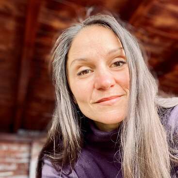 Una mujer con el pelo largo y liso y una sonrisa amable lleva un jersey oscuro de cuello alto. Está en un interior con cálidas vigas de madera de fondo.